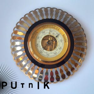 May include: A circular wall-mounted barometer featuring a gold-toned metallic sunburst frame with scalloped edges. The central dial is gold with black text reading REGEN, VERÄNDERLICH, and SCHON, surrounding a visible internal mechanism with two black needles.