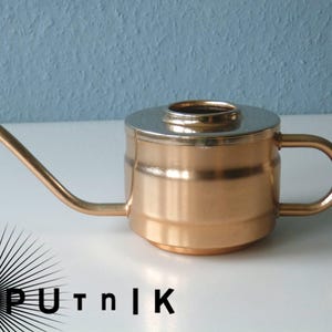 Könnte beinhalten: Eine Gießkanne aus Kupfer mit einem silbernen Deckel und einem Ausguss. Die Gießkanne hat einen Griff an der Seite.
