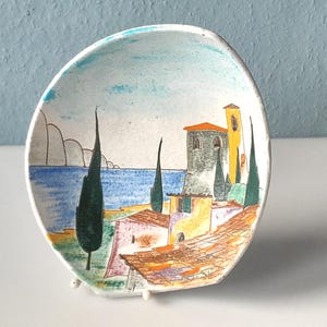 Puede incluir: Un pequeño plato ovalado de cerámica con una escena costera pintada a mano. El diseño presenta un mar azul, un cielo azul claro y edificios coloridos con tejados naranjas. Altos árboles verde oscuro y una torre amarilla completan el paisaje.