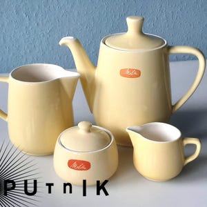 Può includere: Un servizio da tè vintage in ceramica gialla, composto da una teiera, una lattiera, una zuccheriera e un piccolo bricco. Ogni pezzo ha un interno color crema e un'etichetta arancione "Melitta".