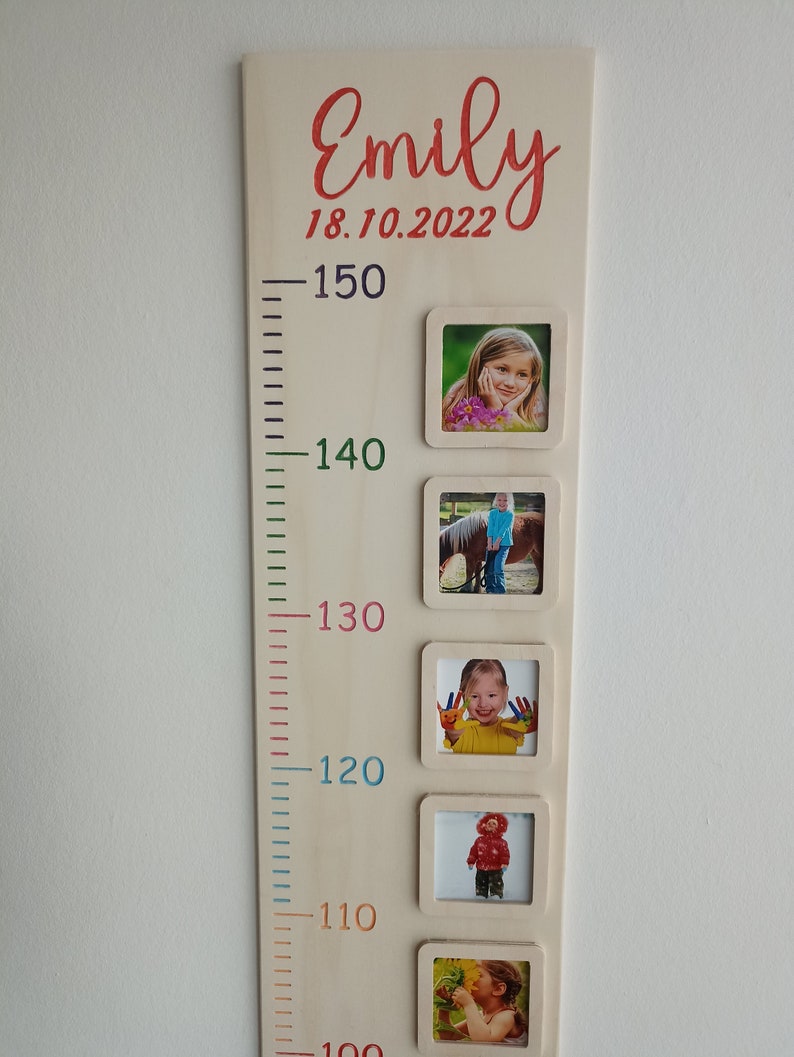 May include: A wooden growth chart with a white background and colourful numbers. The chart has a name, "Emily", and a date, "18.10.2022", written in red. There are five photo frames with pictures of a child at different ages.