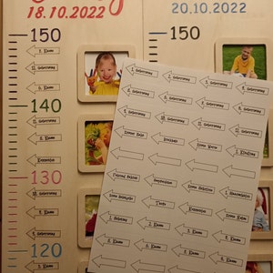 May include: A wooden growth chart with two sections, one for Emily and one for Timo. Each section has a ruler with measurements in centimetres, a photo frame, and a series of stickers with milestones and ages. The stickers are arranged in a row, with arrows pointing to the right. The stickers include milestones such as "First Birthday", "First Steps", "First Tooth", "Kindergarten", and "First Grade".