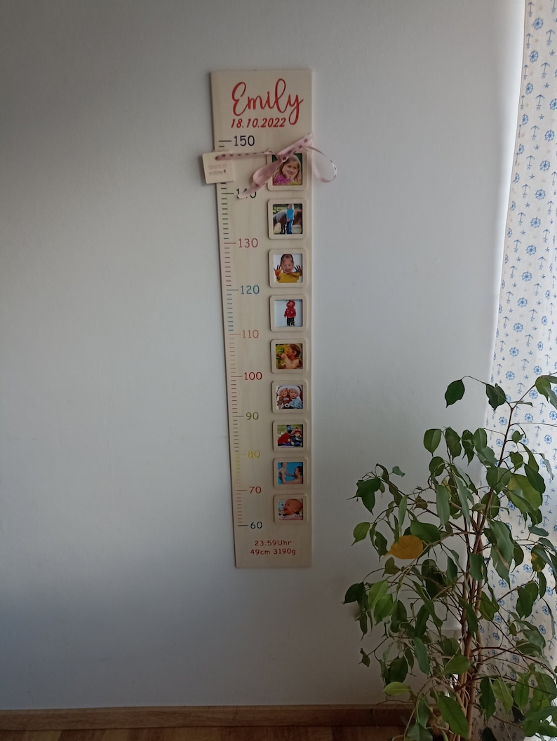 Pode incluir: Um gr&aacute;fico de crescimento de madeira com uma fita rosa e fotos de uma crian&ccedil;a em diferentes idades. O gr&aacute;fico &eacute; etiquetado com o nome "Emily" e a data "18.10.2022". O gr&aacute;fico tamb&eacute;m inclui medidas em polegadas e cent&iacute;metros.