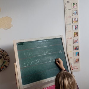 May include: A wooden growth chart with a white background and a red inscription that reads "Emily 18.10.2022". The chart has a ruler with measurements in centimetres from 50 to 150. There are small photo frames with pictures of a child at different ages.