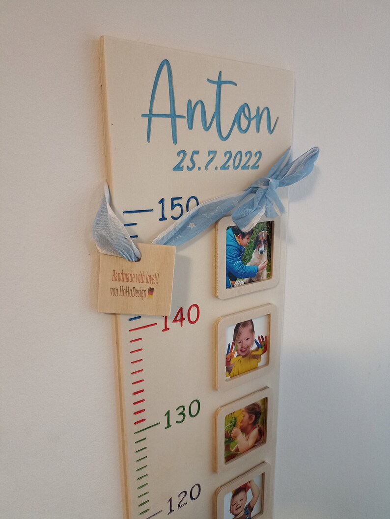 Pode incluir: Um gr&aacute;fico de crescimento de madeira com uma fita azul e branca, uma pequena etiqueta que diz "Handmade with love!! von HoHoDesign", e uma r&eacute;gua com marcas de 120 a 150 cent&iacute;metros. O gr&aacute;fico tem quatro molduras de fotos com imagens de uma crian&ccedil;a em diferentes idades. O gr&aacute;fico est&aacute; etiquetado como "Anton 25.7.2022".