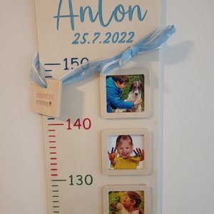 May include: A white wooden growth chart with a blue ribbon, a blue star pattern, and a small wooden tag with the text "Handmade with love by Babble Design". The chart has a ruler with red, green, and blue markings and the numbers 150, 140, 130, and 120. The chart also has four photo frames with pictures of a child. The chart has the name "Anton" and the date "25.7.2022" written in blue.