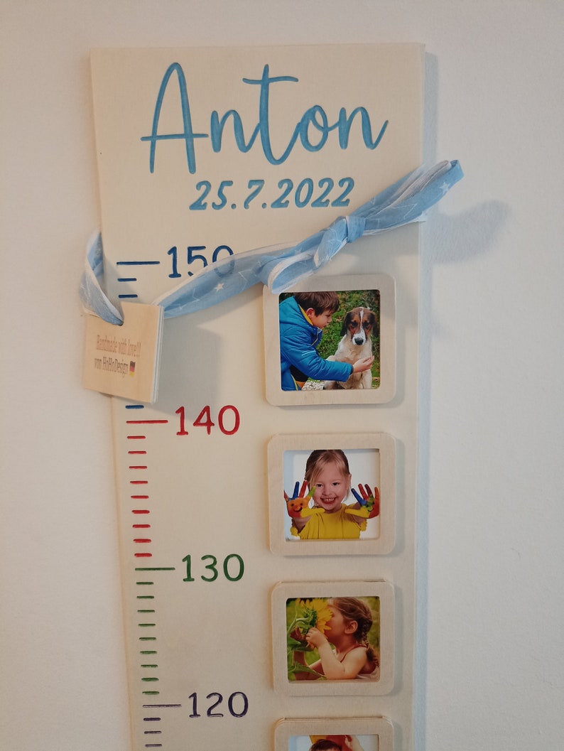 May include: A white wooden growth chart with a blue ribbon, a blue star pattern, and a small wooden tag with the text "Handmade with love by Babble Design". The chart has a ruler with red, green, and blue markings and the numbers 150, 140, 130, and 120. The chart also has four photo frames with pictures of a child. The chart has the name "Anton" and the date "25.7.2022" written in blue.