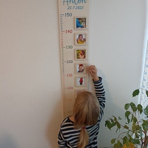 Op de afbeelding: Een houten groeimeter met een witte achtergrond en rode markeringen. De meter heeft een blauwe inscriptie met de tekst "Anton 25.7.2022". Er zijn vijf fotolijsten met foto's van een kind op verschillende leeftijden. Een kind staat naast de meter en reikt omhoog om de bovenste lijst aan te raken.