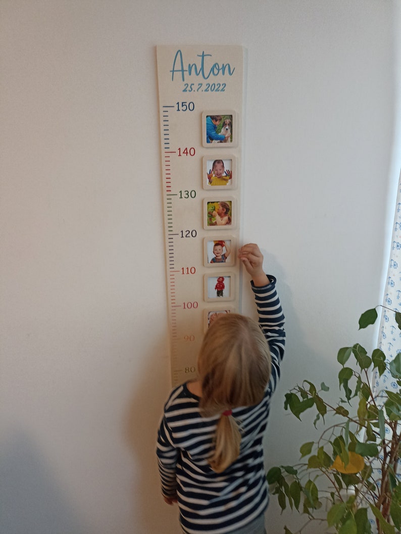 Op de afbeelding: Een houten groeimeter met een witte achtergrond en rode markeringen. De meter heeft een blauwe inscriptie met de tekst "Anton 25.7.2022". Er zijn vijf fotolijsten met foto's van een kind op verschillende leeftijden. Een kind staat naast de meter en reikt omhoog om de bovenste lijst aan te raken.