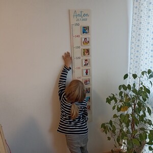 May include: A wooden growth chart with a light brown background and red numbers. The chart is decorated with framed photos of a child at different ages. The chart is labelled "Anton 25.7.2022" at the top. A child is standing next to the chart, reaching up to the 100 cm mark.