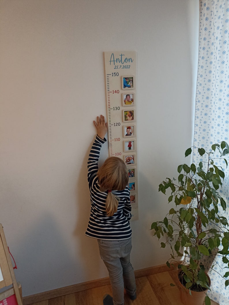 May include: A wooden growth chart with a light brown background and red numbers. The chart is decorated with framed photos of a child at different ages. The chart is labelled "Anton 25.7.2022" at the top. A child is standing next to the chart, reaching up to the 100 cm mark.