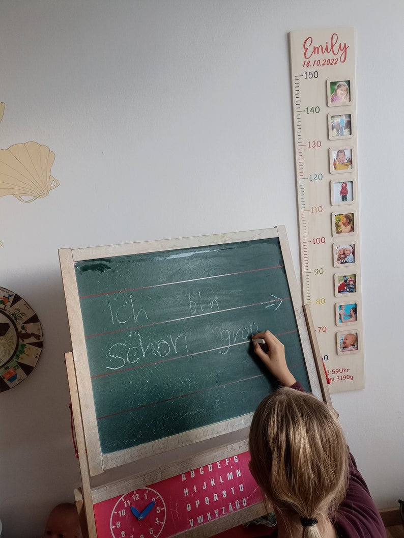 Pode incluir: Um gr&aacute;fico de crescimento de madeira com fundo branco e uma inscri&ccedil;&atilde;o vermelha que diz "Emily 18.10.2022". O gr&aacute;fico tem uma r&eacute;gua com medidas em cent&iacute;metros de 50 a 150. H&aacute; pequenos porta-retratos com fotos de uma crian&ccedil;a em diferentes idades.