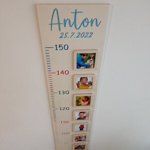 May include: A white wooden growth chart with a blue inscription "Anton 25.7.2022". The chart has a ruler with markings from 70 to 150 cm and photo frames at each height marking. Each frame contains a photo of a child at different ages.