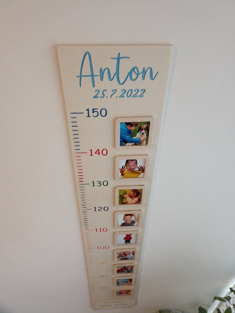 May include: A white wooden growth chart with a blue inscription "Anton 25.7.2022". The chart has a ruler with markings from 70 to 150 cm and photo frames at each height marking. Each frame contains a photo of a child at different ages.