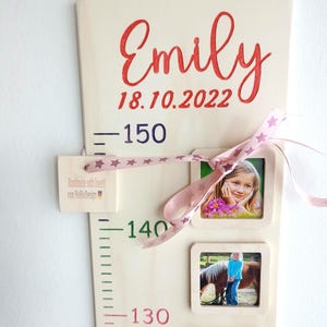 Op de afbeelding: Een houten groeimeter met een roze lint en sterren, met een naam, een datum en hoogtemetingen. De meter heeft vier fotolijstjes met foto's van een kind op verschillende leeftijden. De tekst op de meter luidt "Emily 18.10.2022 - 150 - 140 - 130".