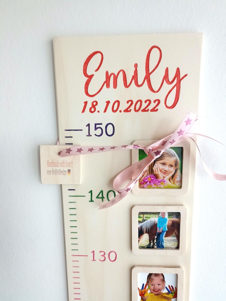Op de afbeelding: Een houten groeimeter met een roze lint en sterren, met een naam, een datum en hoogtemetingen. De meter heeft vier fotolijstjes met foto's van een kind op verschillende leeftijden. De tekst op de meter luidt "Emily 18.10.2022 - 150 - 140 - 130".