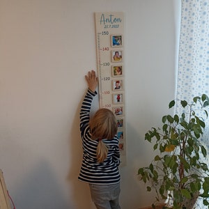 Op de afbeelding: Een houten groeimeter met een lichtbruine achtergrond en rode cijfers. De meter is versierd met ingelijste foto's van een kind op verschillende leeftijden. De meter is bovenaan gelabeld met "Anton 25.7.2022". Een kind staat naast de meter en reikt tot de 100 cm markering.