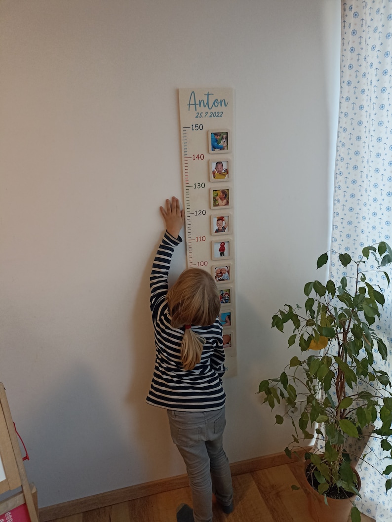 Op de afbeelding: Een houten groeimeter met een lichtbruine achtergrond en rode cijfers. De meter is versierd met ingelijste foto's van een kind op verschillende leeftijden. De meter is bovenaan gelabeld met "Anton 25.7.2022". Een kind staat naast de meter en reikt tot de 100 cm markering.