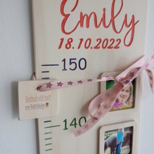 May include: A wooden growth chart with a pink ribbon and stars, featuring the name "Emily" and the date "18.10.2022". The chart has measurements in centimetres, with a small tag that reads "Handmade with love!! von HoHoDesign".