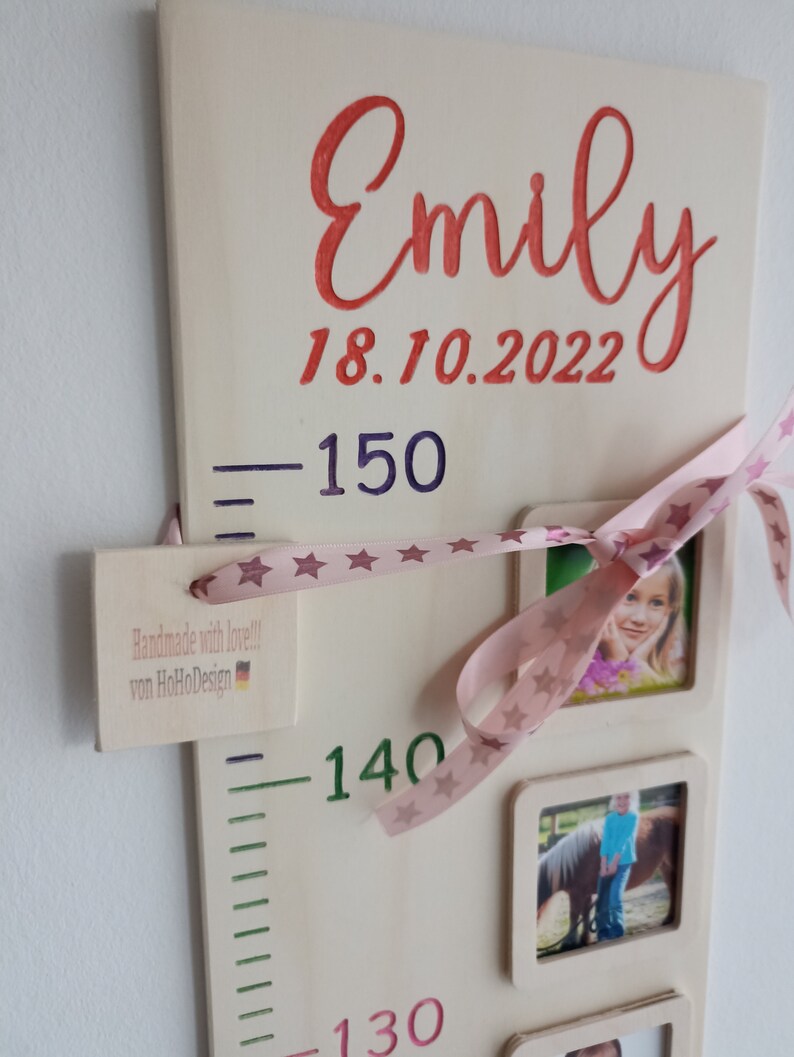 May include: A wooden growth chart with a pink ribbon and stars, featuring the name "Emily" and the date "18.10.2022". The chart has measurements in centimetres, with a small tag that reads "Handmade with love!! von HoHoDesign".