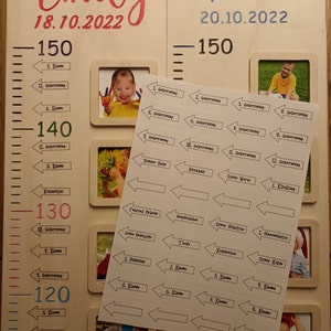 Op de afbeelding: Een witte houten groeimeter met twee secties, een voor Emily en een voor Timo. Elke sectie heeft een liniaal met markeringen voor de hoogte in centimeters. De kaart bevat ook een fotolijst met een foto van een kind en een set stickers met pijlen en tekst die mijlpalen aangeven zoals verjaardagen en schooljaren.