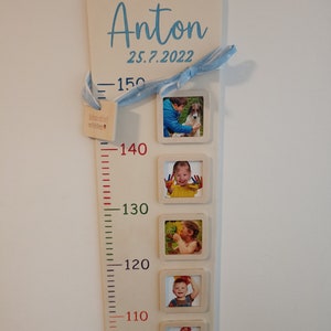 Op de afbeelding: Een witte houten groeimeter met een blauw lint en een blauwe inscriptie die "Anton 25.7.2022" luidt. De meter heeft een liniaal met rode, groene en zwarte markeringen en zes fotolijsten met foto's van een kind op verschillende leeftijden.