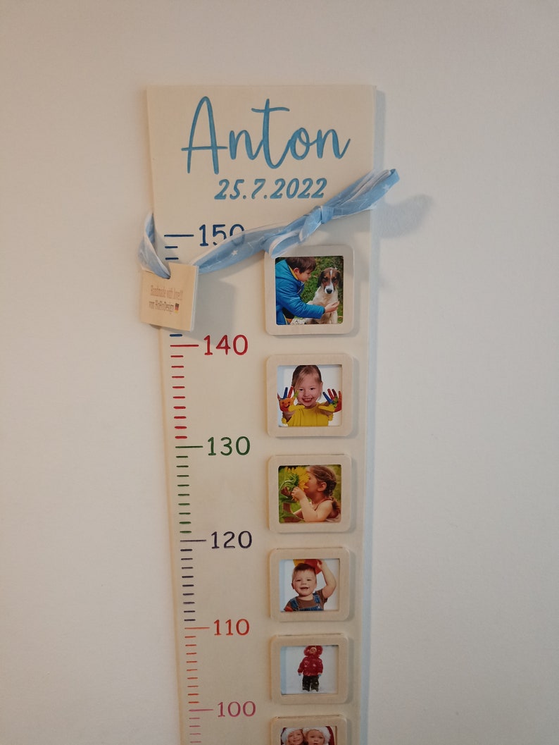 Op de afbeelding: Een witte houten groeimeter met een blauw lint en een blauwe inscriptie die "Anton 25.7.2022" luidt. De meter heeft een liniaal met rode, groene en zwarte markeringen en zes fotolijsten met foto's van een kind op verschillende leeftijden.