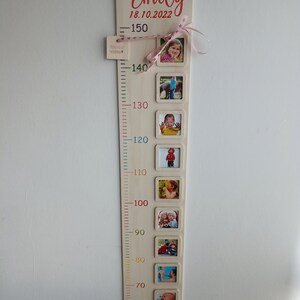 Op de afbeelding: Een houten groeimeter met een roze lint en een naamplaatje met de tekst "Emily 18.10.2022". De meter heeft een schaal van 60 tot 150 centimeter en bevat ruimte voor foto's. De meter bevat ook de tekst "23:59Uhr 49cm 3190g".