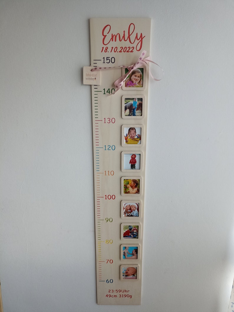 Op de afbeelding: Een houten groeimeter met een roze lint en een naamplaatje met de tekst "Emily 18.10.2022". De meter heeft een schaal van 60 tot 150 centimeter en bevat ruimte voor foto's. De meter bevat ook de tekst "23:59Uhr 49cm 3190g".