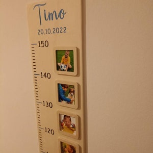 Op de afbeelding: Een houten groeimeter met een witte achtergrond en zwarte markeringen. De meter is gelabeld met "Timo" en "20.10.2022". Er zijn vijf fotolijsten met foto's van een kind op verschillende leeftijden.