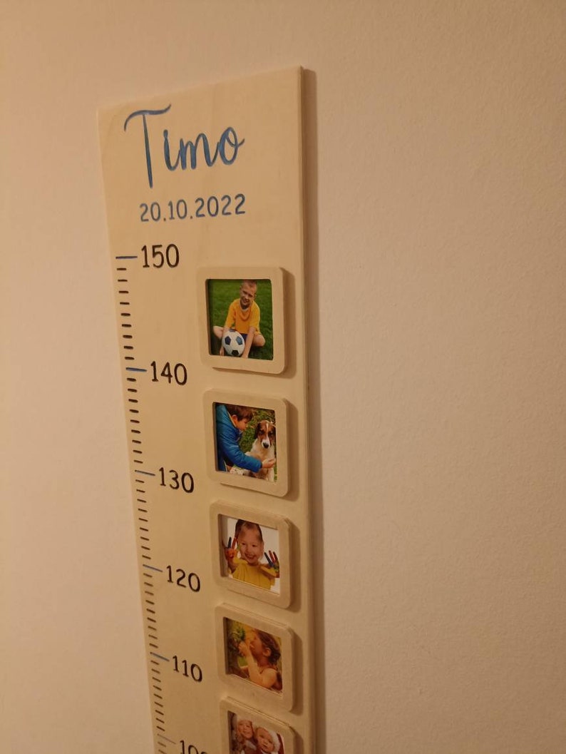 Op de afbeelding: Een houten groeimeter met een witte achtergrond en zwarte markeringen. De meter is gelabeld met "Timo" en "20.10.2022". Er zijn vijf fotolijsten met foto's van een kind op verschillende leeftijden.
