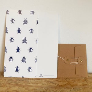 Puede incluir: Una tarjeta blanca con un patrón de escarabajos azules y el texto "The Addressing Joy" impreso en la esquina inferior derecha. La tarjeta está junto a una tarjeta blanca en blanco y un sobre marrón con dos ojales de latón y una cuerda blanca.