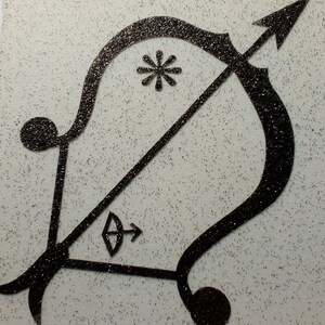 Peut inclure: Un design d'arc et de flèche en paillettes noires sur un fond blanc. La flèche pointe vers la droite et l'arc est courbé vers la gauche. Il y a un motif d'étoile éclatante au centre de l'arc.