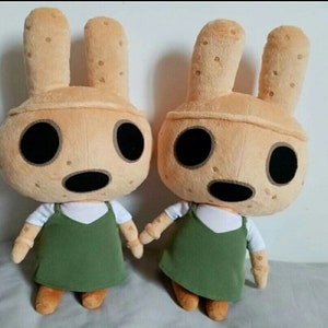 Pu&ograve; includere: Due peluche di un coniglio marrone chiaro con una camicia bianca e un vestito verde. Il coniglio ha gli occhi neri e un naso nero.