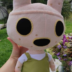 Pu&ograve; includere: Un coniglio di peluche rosa con occhi neri e bocca nera. Il coniglio indossa una maglietta bianca e un vestito con bretelle verde.