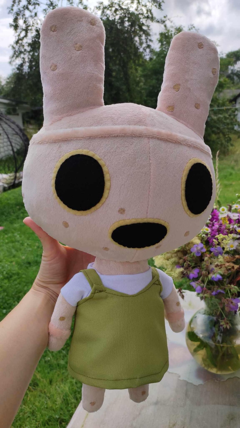 Pu&ograve; includere: Un coniglio di peluche rosa con occhi neri e bocca nera. Il coniglio indossa una maglietta bianca e un vestito con bretelle verde.