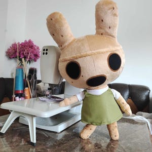 Pu&ograve; includere: Un giocattolo di peluche di un animale marrone chiaro con grandi orecchie e occhi neri. Il giocattolo indossa una camicia bianca e un vestito verde. Il giocattolo &egrave; in piedi accanto a una macchina da cucire bianca.