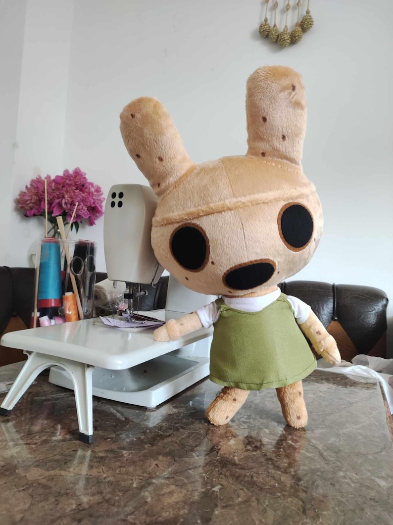 Pu&ograve; includere: Un giocattolo di peluche di un animale marrone chiaro con grandi orecchie e occhi neri. Il giocattolo indossa una camicia bianca e un vestito verde. Il giocattolo &egrave; in piedi accanto a una macchina da cucire bianca.