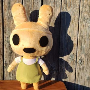 Pu&ograve; includere: Un giocattolo di peluche a forma di coniglio marrone chiaro con grandi occhi neri e un vestito verde. Il coniglio ha orecchie lunghe e un piccolo naso nero.
