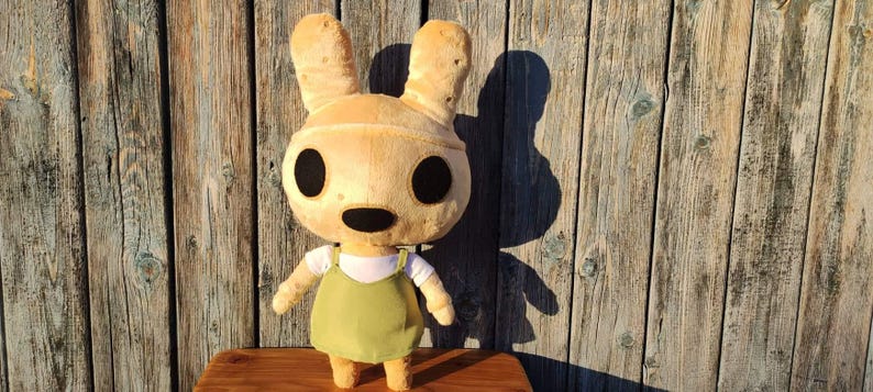 Pu&ograve; includere: Un giocattolo di peluche a forma di coniglio marrone chiaro con grandi occhi neri e un vestito verde. Il coniglio ha orecchie lunghe e un piccolo naso nero.