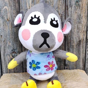 Pode incluir: Um macaco de pelúcia cinza e branco com uma camisa amarela e sapatos amarelos. O macaco tem um nariz rosa e olhos pretos grandes.