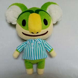 May include: Green and yellow plush toy with white ears and a blue and white striped shirt. The toy has a smiling face and a large yellow beak.