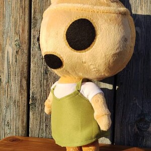 Pu&ograve; includere: Un giocattolo di peluche di un coniglio marrone chiaro che indossa una camicia bianca e un vestito verde. Il coniglio ha grandi occhi neri e una consistenza soffice.