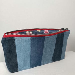 Könnte beinhalten: Eine Jeans-Reißverschlusstasche mit einem gestreiften Design in verschiedenen Blautönen. Das Innenfutter hat ein Blumenmuster. Der Reißverschluss ist silberfarben und die Innenverkleidung ist rot. Diese Tasche eignet sich zur Aufbewahrung von Kosmetika oder kleinen Gegenständen.