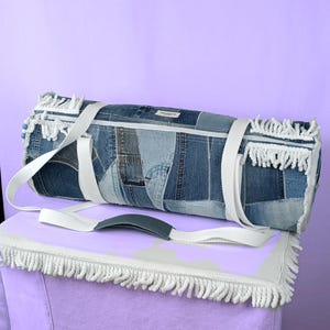 Könnte beinhalten: Eine Jeans-Duffel-Tasche mit weißen Trägern und Fransen. Die Tasche besteht aus Patchwork-Denim in verschiedenen Blautönen. Die Tasche hat einen weißen Riemen mit einem grauen Griff zum Tragen. Die Tasche steht auf einer lilafarbenen Oberfläche.