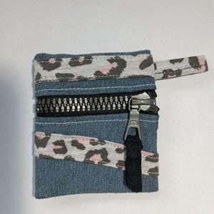 Op de afbeelding: Kleine denim portemonnee met een zwarte ritssluiting en een luipaardprint accent in roze, grijs en wit. De portemonnee heeft een kleine stoffen lus bevestigd aan de ritssluiting.
