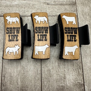 Puede incluir: Tres clips de madera con las palabras "SHOW LIFE" en letras negras. Cada clip tiene una silueta de animal blanco encima del texto. Los clips están sobre una superficie de madera.