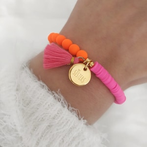 Pode incluir: Uma pulseira com contas laranja e rosa, uma borla rosa e um berloque dourado com a palavra "Sisters". Um acessório vibrante e colorido.