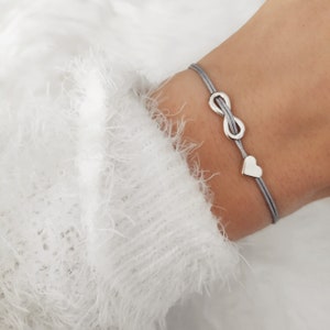 Puede incluir: Pulsera plateada con un símbolo de infinito y un corazón en un cordón gris. La pulsera se lleva en la muñeca, con un suéter blanco en el fondo. El símbolo del infinito está en el centro, y el corazón en un lado.