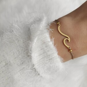 Könnte beinhalten: Goldfarbenes Wellenarmband an einem beigefarbenen Kordelband. Das Wellendesign ist eine einfache, elegante Form. Das Armband wird am Handgelenk getragen, vor einem weißen, flauschigen Pullover.